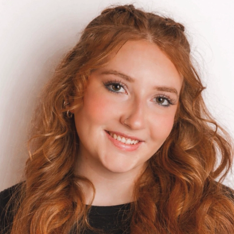 A young woman with long, wavy auburn hair smiles at the camera. She has fair skin, light makeup, and wears a black top. Her hair is parted in the middle and styled with loose curls. The background is plain white.