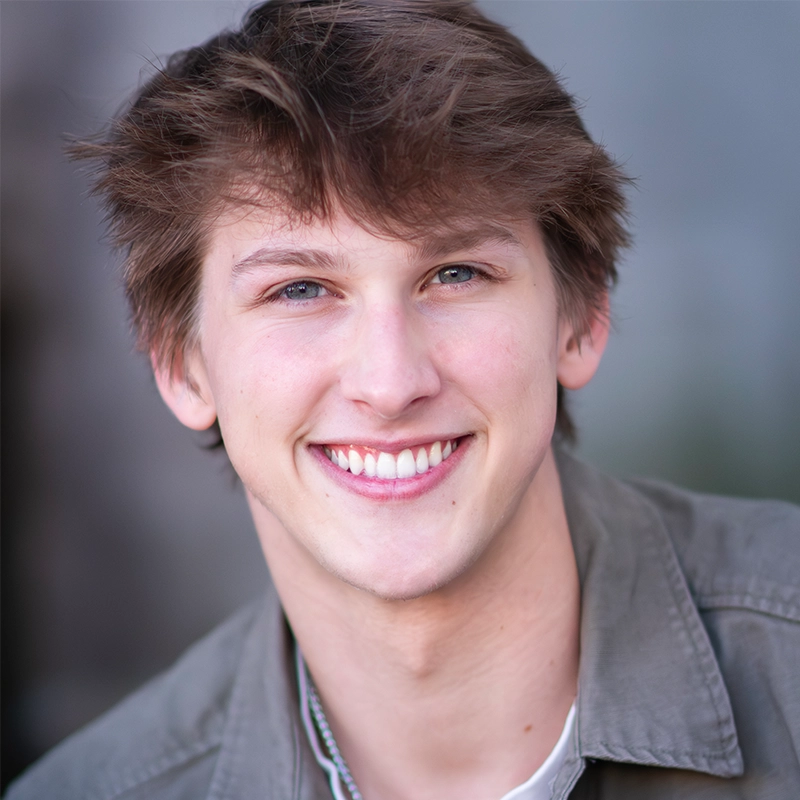 A close-up portrait of Ryan Bagnolati-Camargo, a smiling young man with light brown hair and blue eyes. He has a fair complexion and wears an olive-green collared button-down shirt over a white undershirt against a soft, neutral, out-of-focus background.