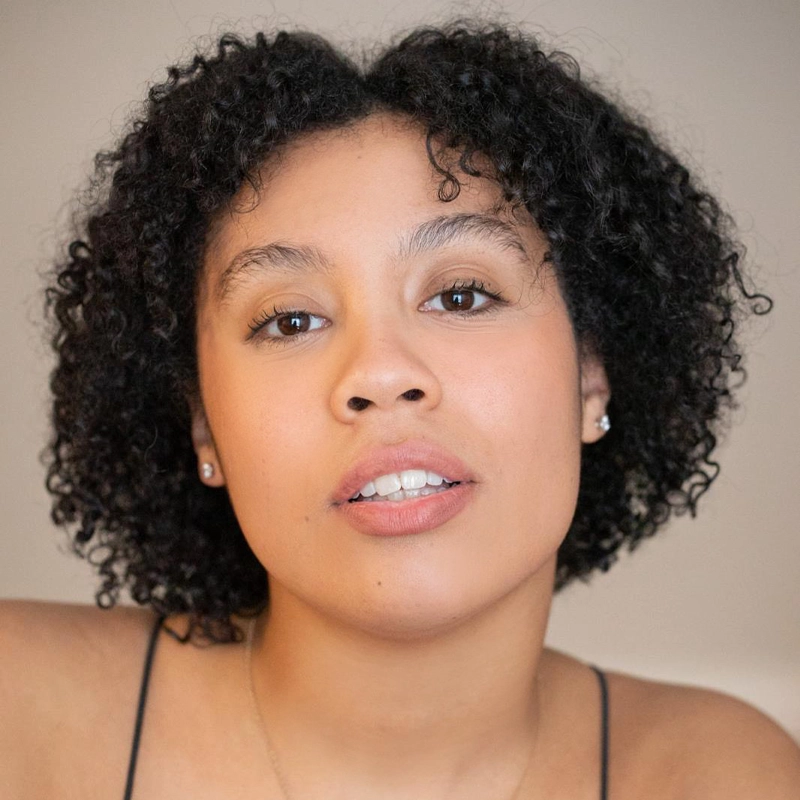 A woman with short curly hair and glowing skin looks directly at the camera. She has light makeup, wears small stud earrings, and a black spaghetti strap top. The softly lit background highlights Sawyer Evans as the focal point of the image.