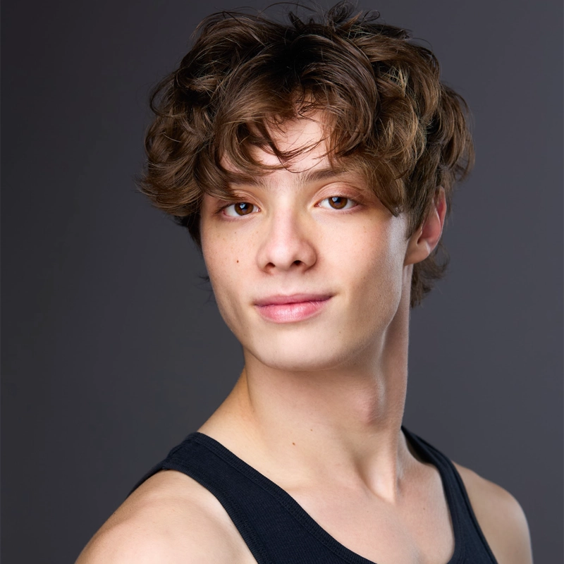 Young person with wavy brown hair, fair skin, and brown eyes, wearing a black tank top. Reilly Favacho faces the camera with a slight smile against a plain, softly-lit gray background that highlights their features and hair texture.