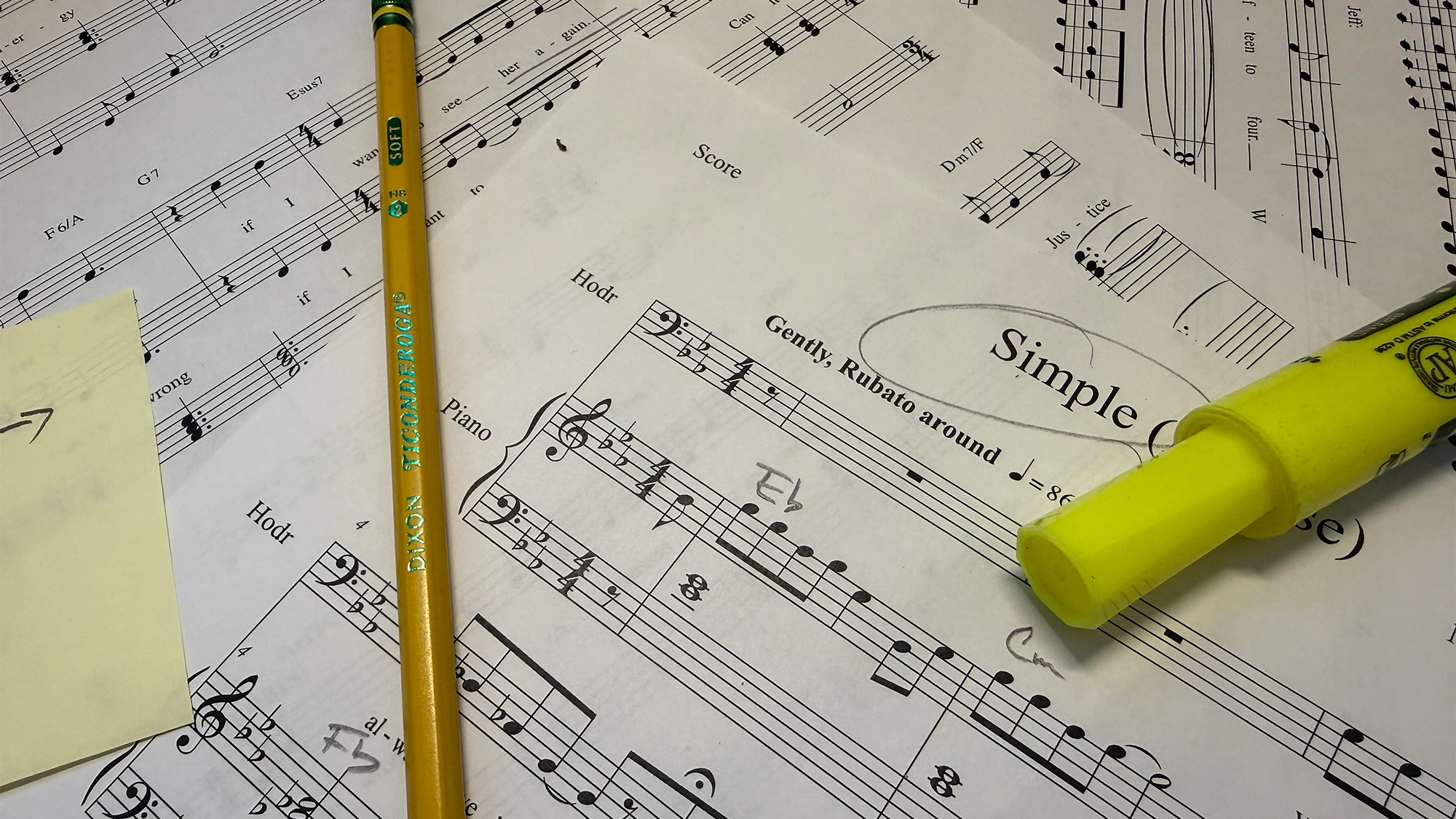 A yellow pencil and a yellow highlighter rest on sheets of musical theatre scores. One page features a circled title reading “Simple” and handwritten musical notes, while a yellow sticky note is partially visible on these first drafts.