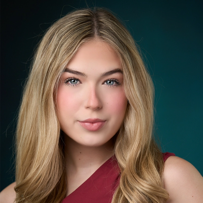 A headshot of Ryan Bagnolati-Camargo, a young woman with long, blonde hair, bright blue eyes, and a neutral expression. She wears a maroon-colored top against a dark, teal-toned background that transitions from black to a deep blue-green.