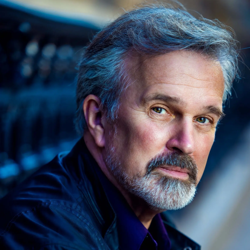 A middle-aged man with graying hair, a mustache, and a goatee looks directly at the camera. He is wearing a dark jacket over a purple shirt.