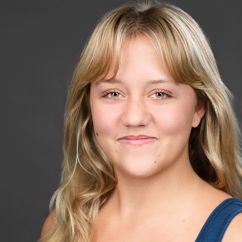 A young woman with long blonde hair and light skin smiles gently at the camera. She wears a sleeveless navy blue top. The background is plain and dark gray, putting focus on her face. Her hair is parted in the middle and falls loosely over her shoulders.