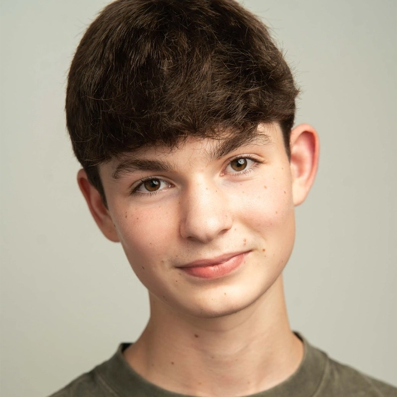 A head-and-shoulders portrait of a young man with short, dark brown hair and a slight smile. He has light skin with faint freckles, brown eyes, and is wearing a dark olive-green crew-neck shirt against a plain, light grey background.