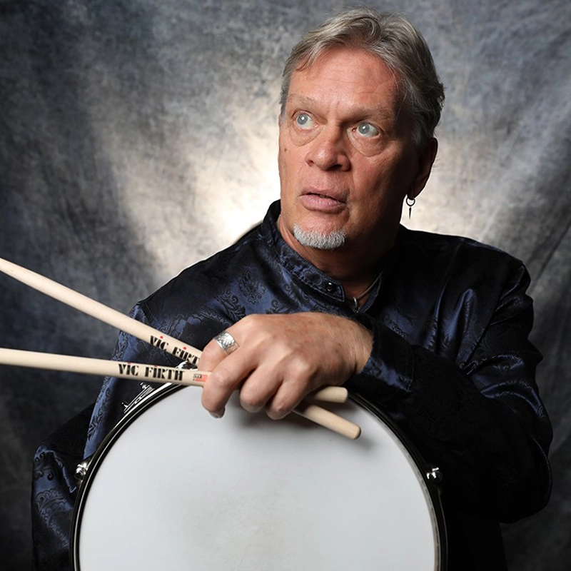 A middle-aged man with short, graying hair and a goatee gazes upward while holding two Vic Firth drumsticks over a white snare drum. He wears a dark, patterned silk shirt and a silver ring. The background is a textured, mottled gray.
