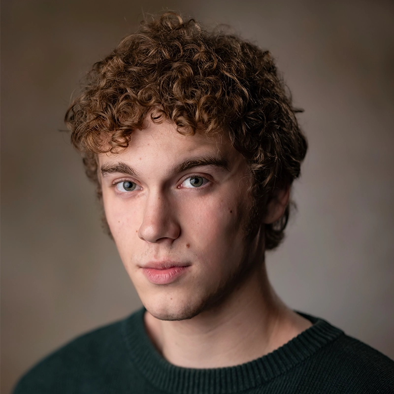 A headshot of Ryan Bagnolati-Camargo, a young man with curly brown hair looking directly at the camera. He has light skin, green eyes, and is wearing a dark green knit sweater against a mottled gray-brown background.