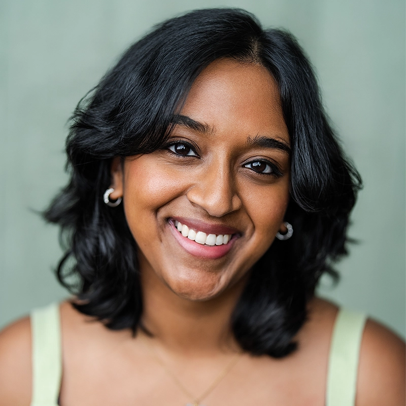 A woman with medium skin, shoulder-length black hair, and silver hoop earrings smiles warmly at the camera. She wears a light green top and a thin necklace, with a soft green background behind her.