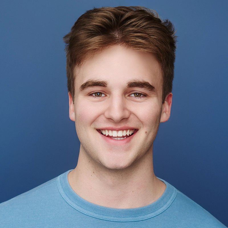 Ryan Bagnolati-Camargo is a young man with light brown hair and hazel eyes who smiles broadly at the camera. He wears a plain light blue crew-neck shirt against a solid, matching blue background.