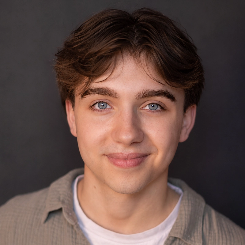 A close-up headshot of Ryan Bagnolati-Camargo, a young man with brown hair, light eyes, and a gentle smile. He is wearing a white crew-neck t-shirt under a light olive-green button-down shirt against a dark, solid background.