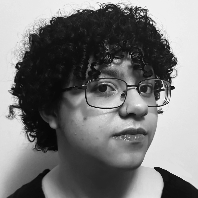 A person with short, curly dark hair and glasses looks at the camera. They have a nose ring and glossy lips, wearing a dark top. The background is plain and light-colored. The photo is black and white.
