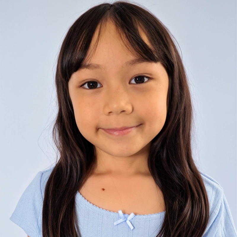 A young girl with long, dark brown hair and bangs smiles gently at the camera. She wears a light blue ribbed top with a small ribbon detail at the neckline against a plain, soft blue background.