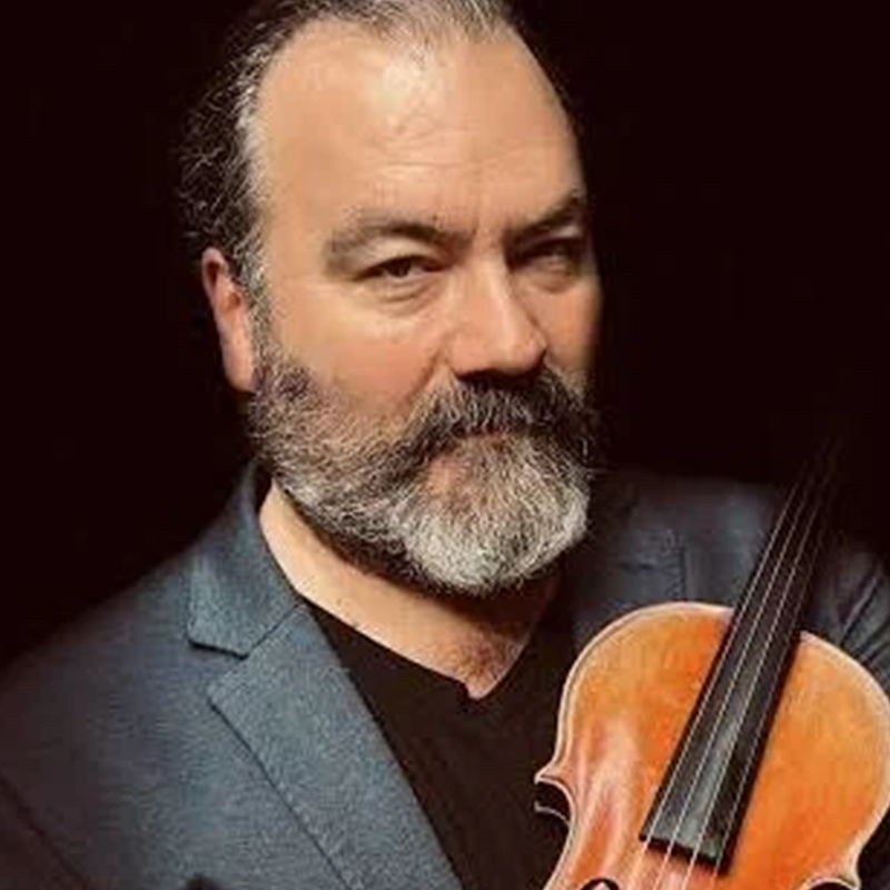 A middle-aged man with a salt-and-pepper beard and hair, wearing a dark blazer and black shirt, holds a violin. The black background highlights Jonathan Dinklage and his instrument as he looks directly at the camera with a neutral expression.