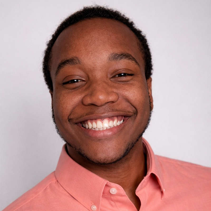 A portrait of Ryan Camargo, a young Black man with a warm smile, wearing a salmon-colored button-down shirt. He is centered against a plain, light gray background, looking directly into the camera.