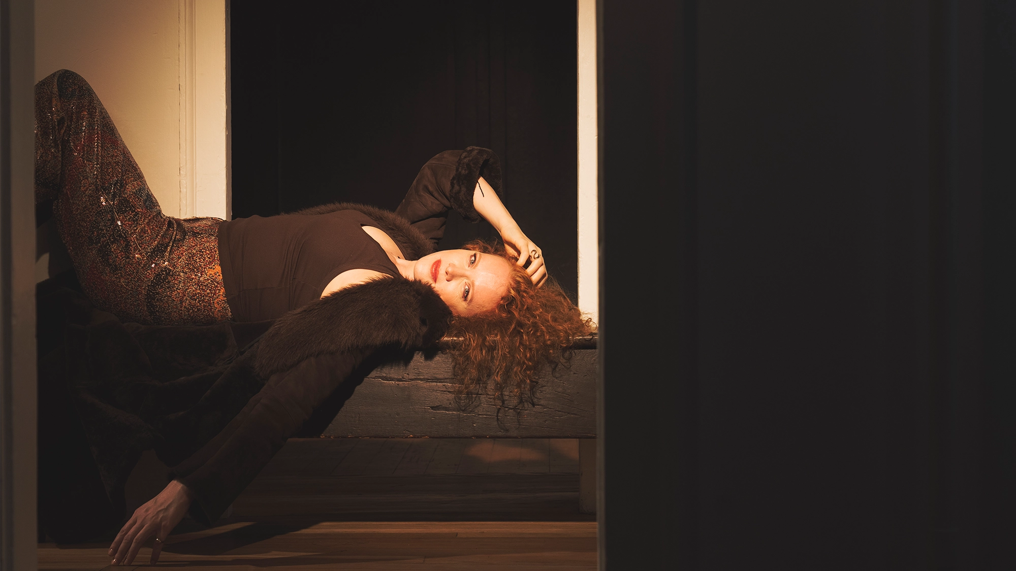 Jazz singer Kristen Lee Sergeant reclines on a dark bench, her curly red hair illuminated by warm light. She wears a black top with fur-trimmed sleeves and shimmery, patterned trousers, gazing toward the camera against the deep shadows of the room.