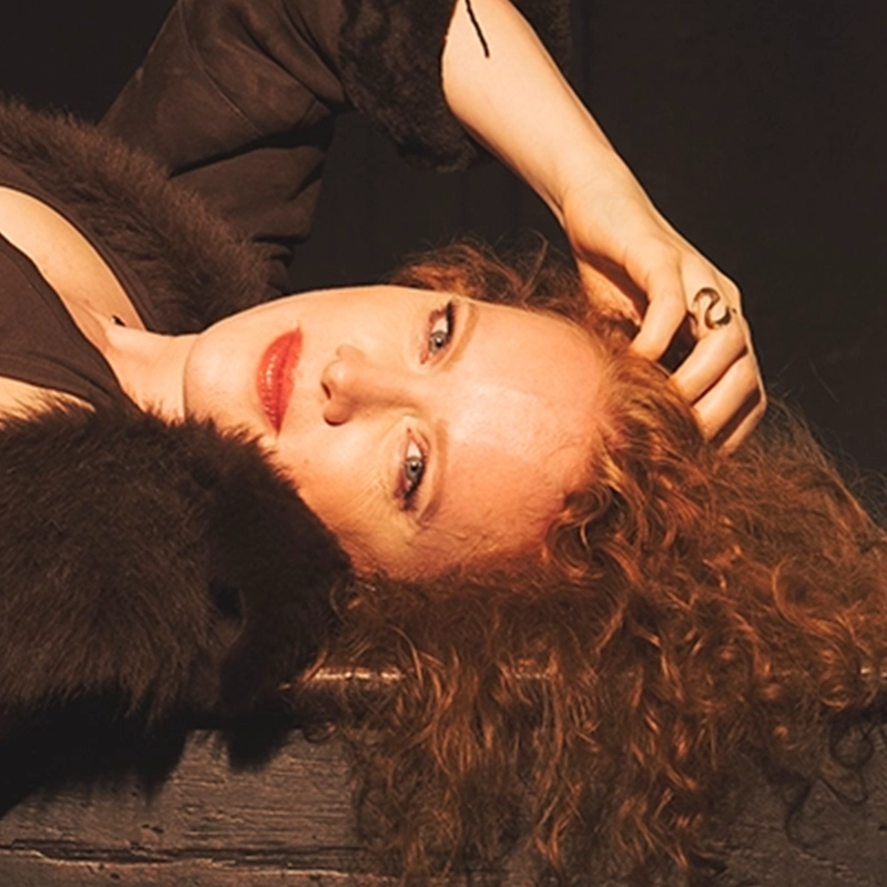Jazz vocalist Kristen Lee Sergeant, with curly auburn hair and red lipstick, lies on a dark surface resting her head on her hand. She wears a black garment with a faux fur trim and a gold ring on her finger, illuminated by warm light against a dark background.