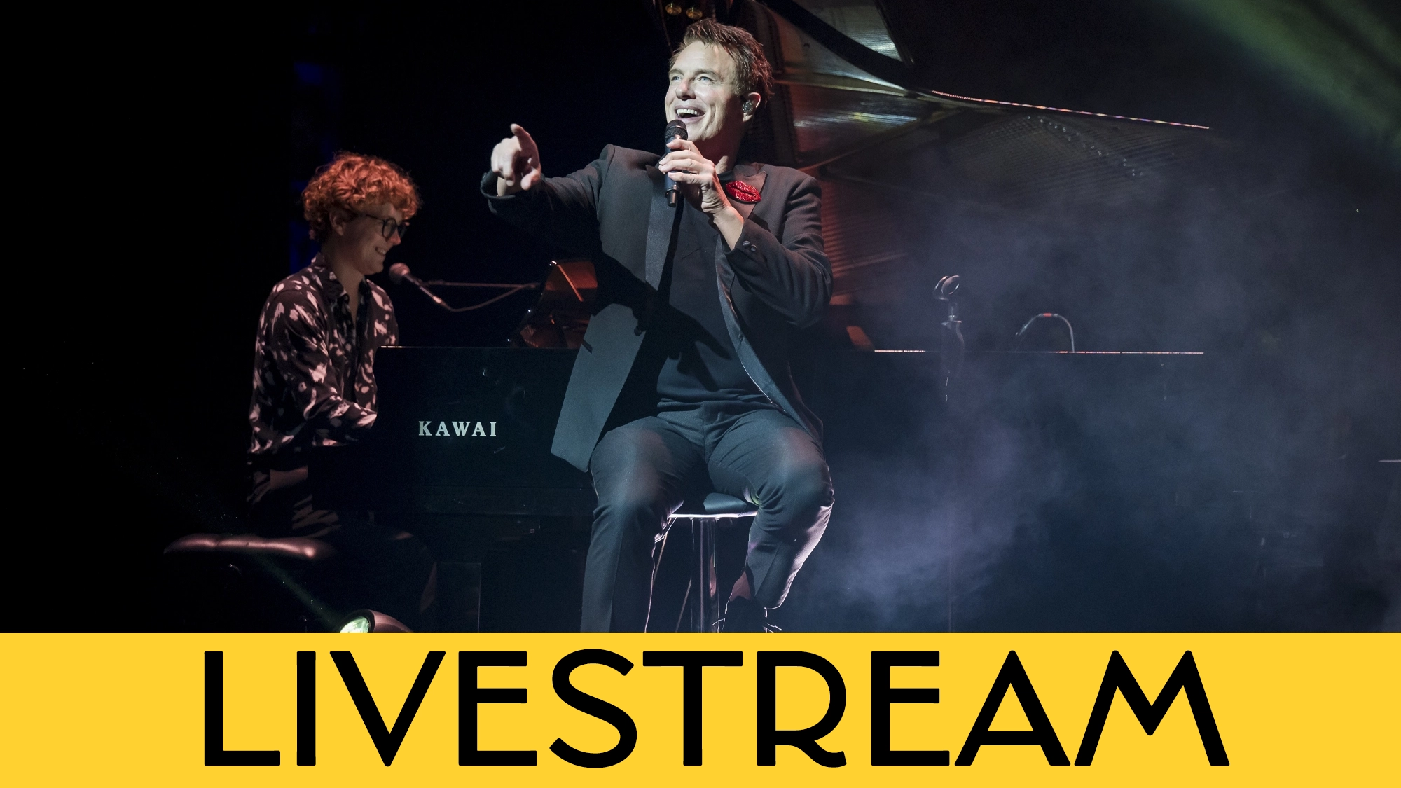John Barrowman in a black suit sits on a stage stool, singing into a microphone and gesturing to the audience. Behind him, a person with curly hair plays a Kawai grand piano in the smoky venue. A bright yellow bar at the bottom contains the black text “LIVESTREAM.”