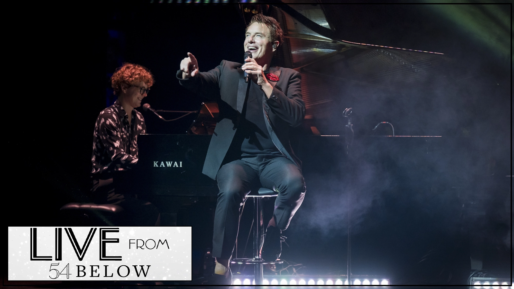 LIVE John Barrowman John Barrowman, wearing a dark suit and red lapel pin, sings into a microphone while gesturing from a stool. Behind him, a person with curly hair plays a Kawai grand piano under moody stage lighting, captured during a celebrity livestream with “LIVE FROM 54 BELOW” in the corner.