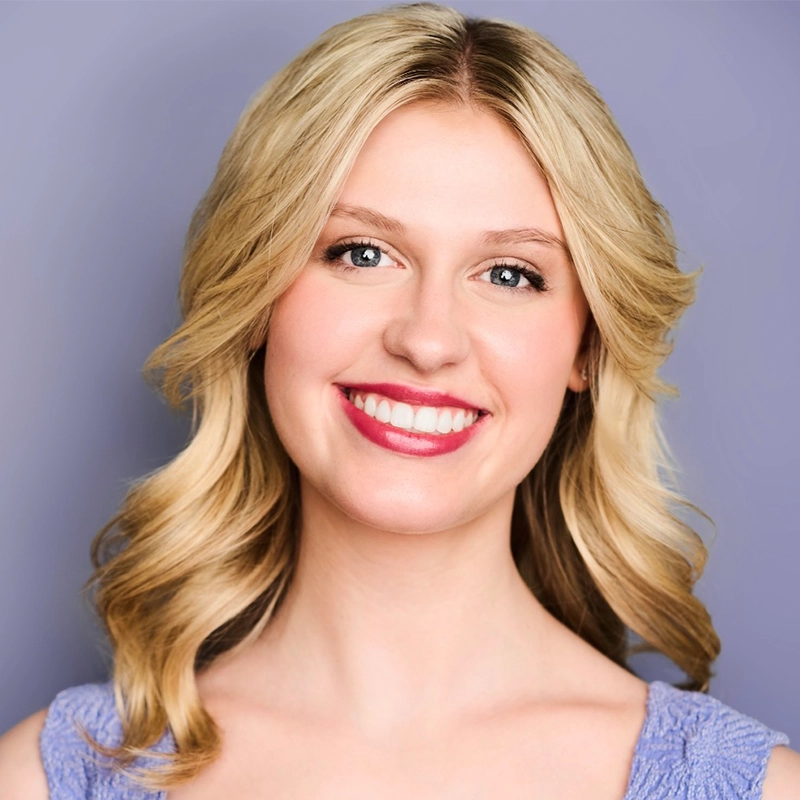 Ryan Bagnolati-Camargo poses for a headshot, smiling brightly with wavy blonde hair. She wears a textured, light purple top against a matching lavender background.