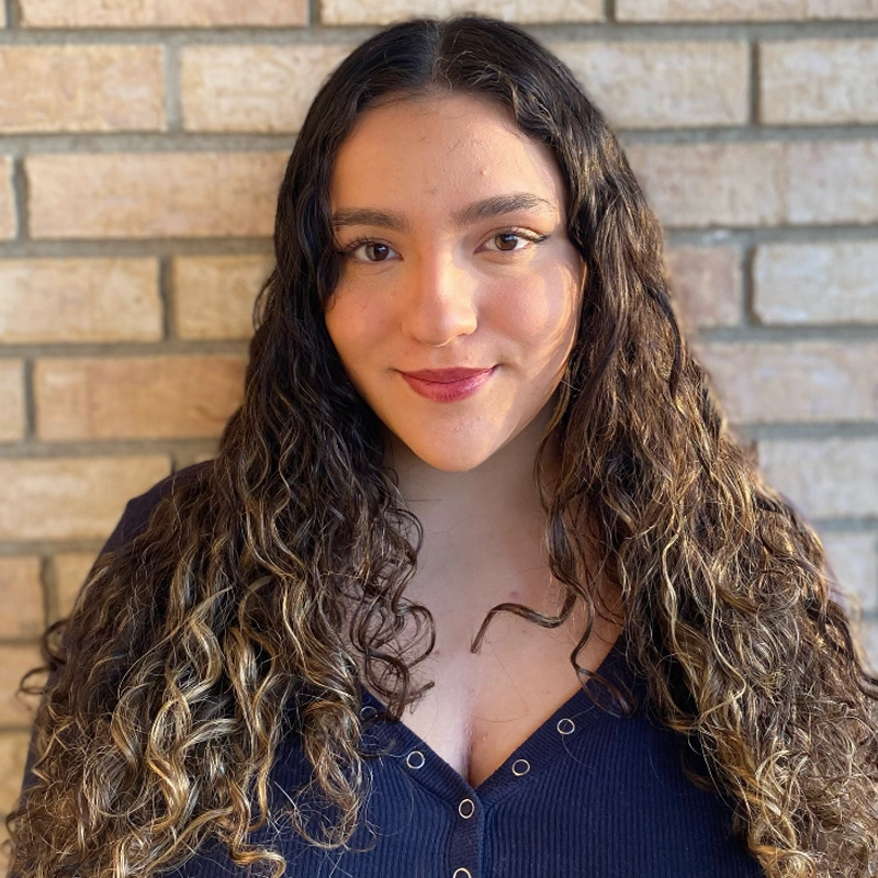 Juliana LoPresti, with long, curly brown hair and highlights, smiles at the camera. She wears a dark, buttoned top. The background is a light brick wall, and sunlight softly illuminates her face and hair.