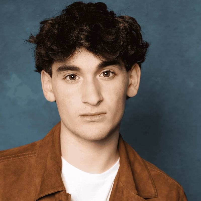A portrait of Justin Lowenhar, a young man with dark, wavy hair, brown eyes, and a neutral expression. He is wearing a white crew-neck t-shirt under a tan suede jacket against a textured blue backdrop.