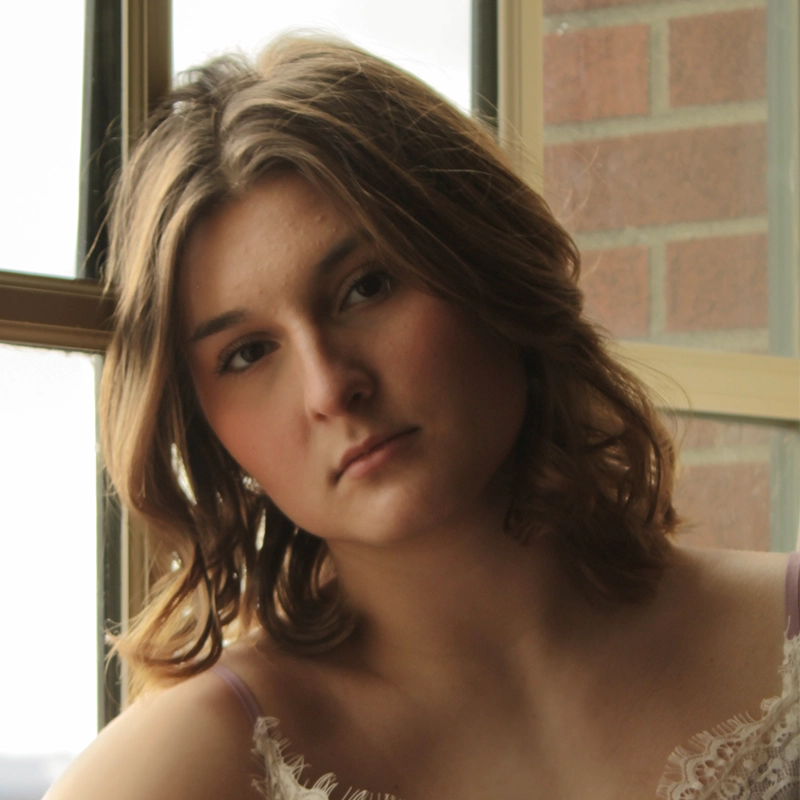 Ryan Bagnolati-Camargo, a young woman with shoulder-length, wavy brown hair, looks directly into the camera. She wears a cream-colored lace top and sits in front of a window with a brick wall visible in the background.