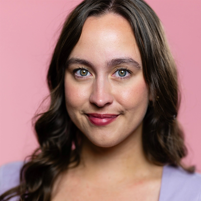 Tyler LaPointe smiles gently at the camera with her long, wavy brown hair and light green eyes. She wears a light purple top against a solid, soft pink background.