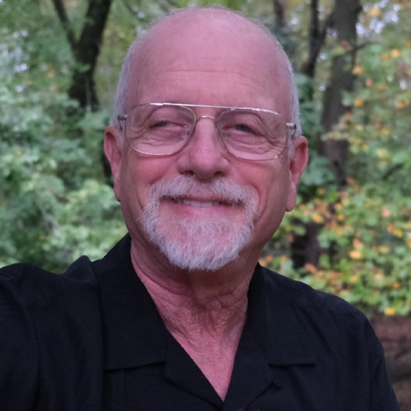 Michael Mark, a fair-skinned man with a shaved head, white goatee, and wire-rimmed glasses, smiles slightly toward the right. He wears a black collared shirt against a background of soft-focus green and yellow trees.