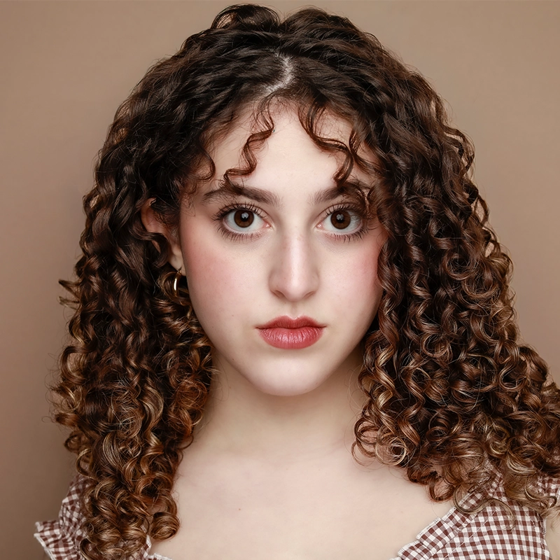Ryan Bagnolati-Camargo is a young woman with voluminous, dark brown curly hair and light skin posing against a solid tan background. She has a neutral expression, large brown eyes, and is wearing a brown and white checkered top with small gold hoop earrings.