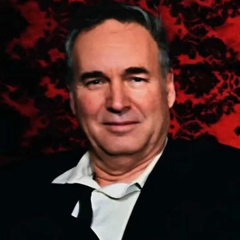 A middle-aged man with short, greying hair and a neutral expression, posing like Charlene Hansen in a white collared shirt and black jacket against a textured, deep red backdrop with a dark, ornate damask-like pattern.