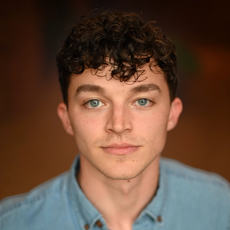 A headshot of Tyler LaPointe, a young man with curly dark hair and light eyes looking directly at the camera. He has a neutral expression and is wearing a light blue button-down shirt against a soft, dark, out-of-focus background.