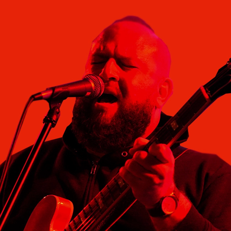A bearded man with a shaved head sings passionately into a microphone while playing an electric guitar. He wears a dark zip-up jacket and a watch, set against a vibrant, solid red background.