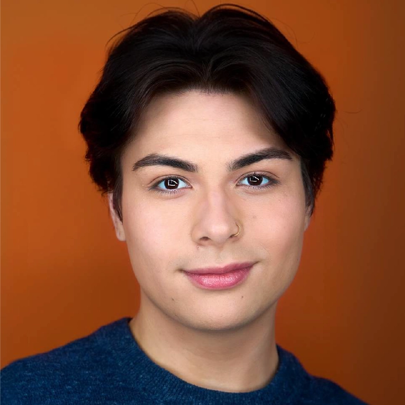 A portrait of a person with dark, center-parted hair, brown eyes, and a small gold nose ring, looking directly at the camera with a gentle smile. They are wearing a textured blue sweater against a solid, warm orange background.