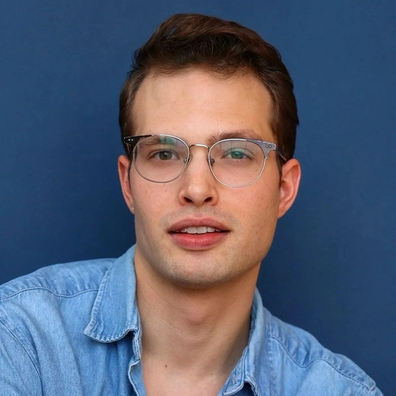 Maxwell Carmel, a young man with short brown hair, wears clear-lens glasses with metal frames and a blue button-down shirt. He is looking at the camera against a solid blue background.