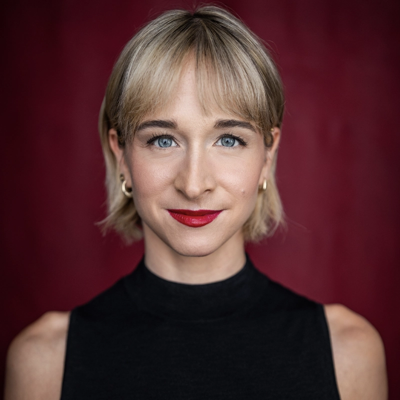 A headshot of a woman with short blonde hair and bright red lipstick. She is wearing a black high-neck top against a solid dark red background.