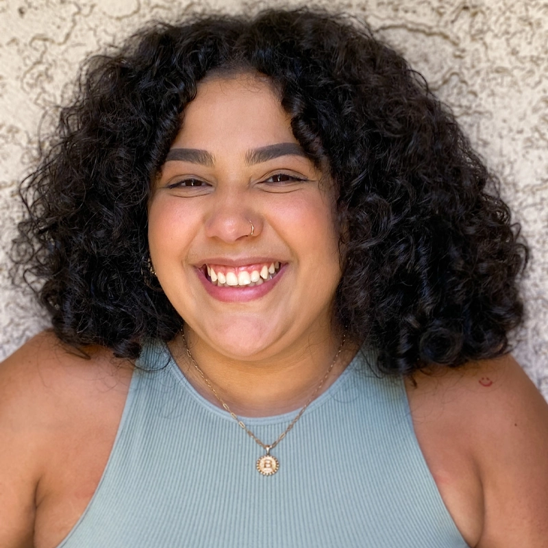 Bryanna Navarro smiles, wearing voluminous curly dark hair and a gold nose ring. They are dressed in a light blue high-neck top and a gold necklace with a circular initial pendant against a textured, off-white wall.