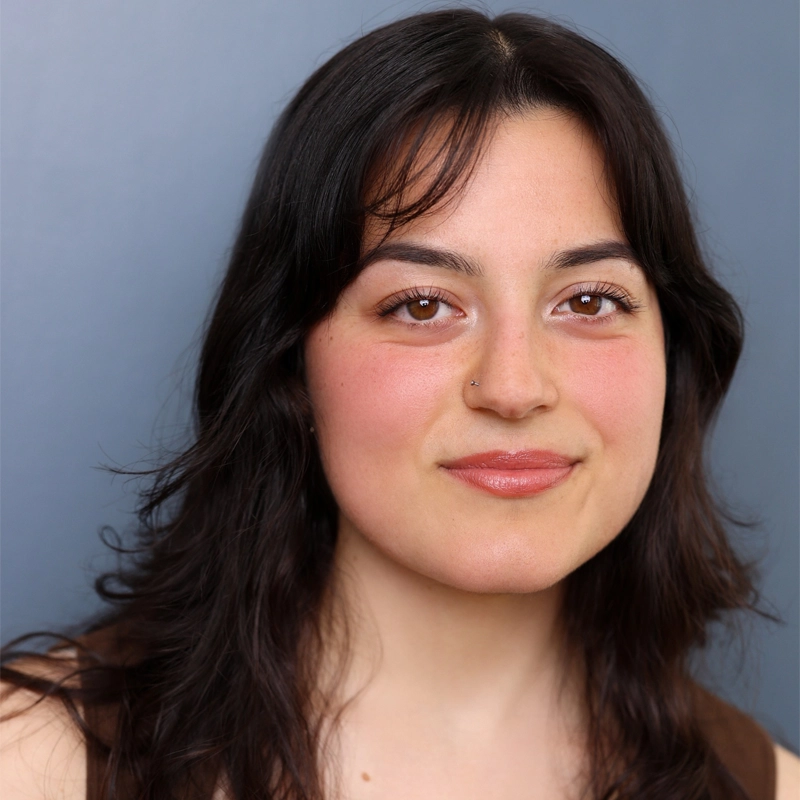 Victoria Nichols, a young woman with medium-length dark brown hair and brown eyes, smiles softly. She has a nose piercing and natural makeup with blush and peach lipstick. The solid gray-blue background highlights her brown top.