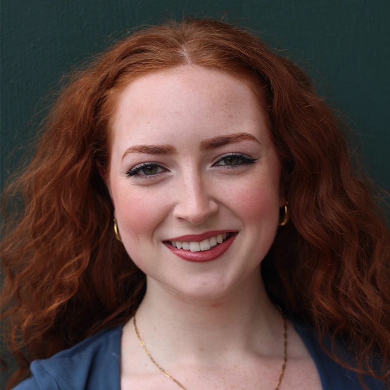 A woman with long curly red hair, fair skin, and blue eyes smiles at the camera. She wears gold hoop earrings, a delicate necklace, natural makeup, and a blue top. The background is a solid dark green color.