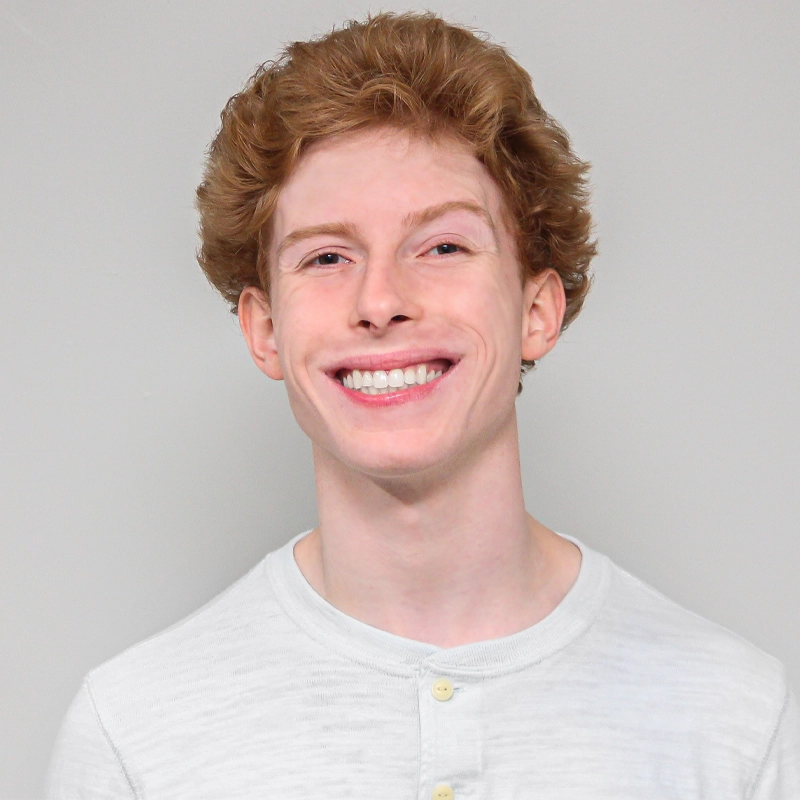 Quinn O’Brien, a young person with fair skin and wavy, light brown hair, is smiling widely and showing their teeth. They are wearing a light gray buttoned shirt against a plain, light-colored background.