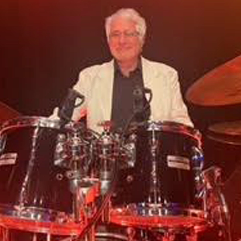 A smiling, gray-haired man in a light-colored blazer and dark shirt plays a drum set. He sits behind two large, glossy black tom-tom drums under warm, reddish stage lighting.