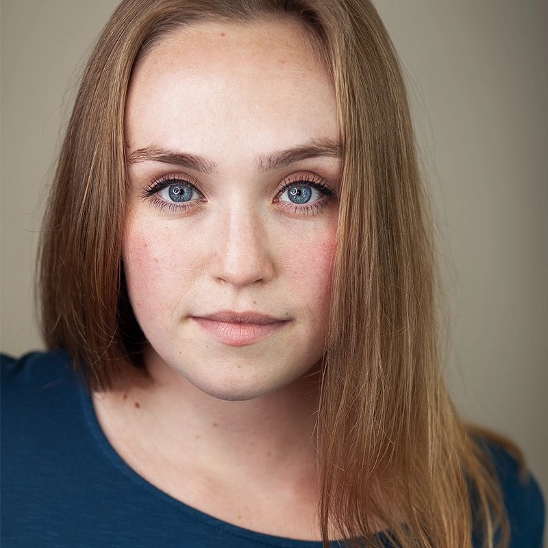 A headshot of Patty Anne Irwin with long, straight light-brown hair, bright blue eyes, and a neutral expression. She wears a plain dark teal shirt against a solid, light beige background.