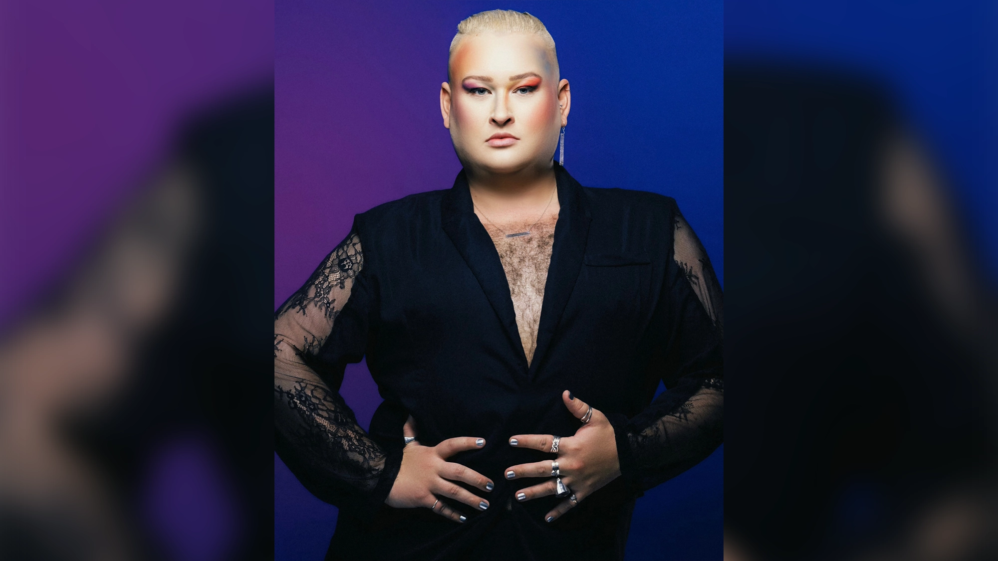 Ryan James Monroe with bleached blonde hair and colorful eye makeup wears a black blazer with lace sleeves and silver rings, posing against a backdrop that gradients from purple to blue.