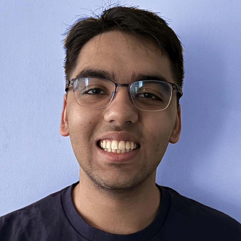 Poorv Ramchandani, a young man with short dark hair and glasses, smiles broadly at the camera. He is wearing a dark t-shirt against a plain, light blue background.