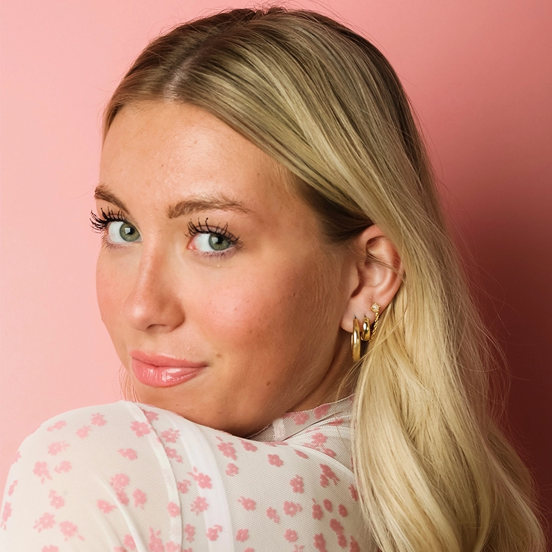 A blonde woman with green eyes, captured by Ryan Bagnolati-Camargo, looks over her shoulder at the camera against a soft pink background. She wears a sheer white top patterned with small pink flowers and small gold hoop earrings.