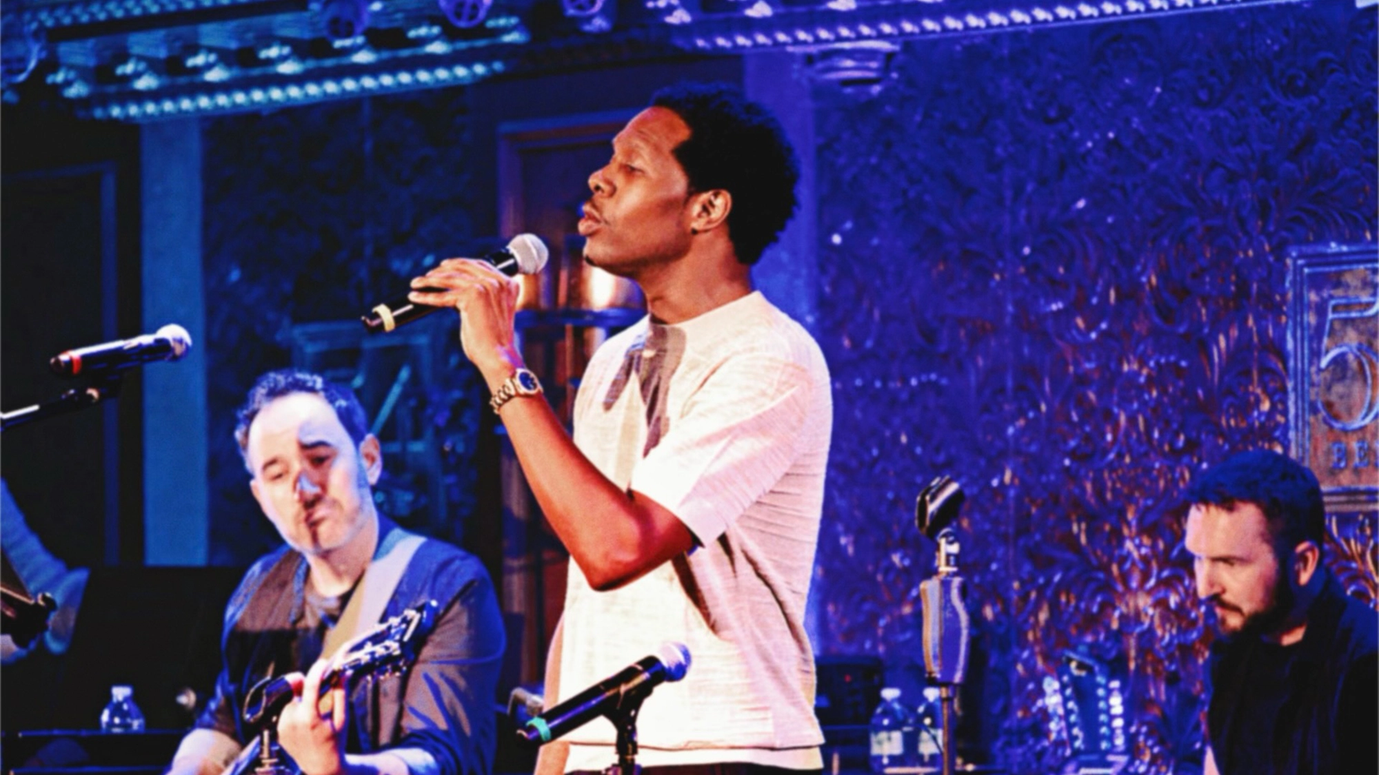Richard Baskin Jr. sings into a microphone on a dimly lit stage, flanked by a guitarist on the left and another musician on the right against a blue, textured backdrop.
