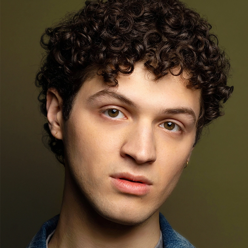 A young person with light skin and short, dark curly hair looks at the camera with a neutral expression. They wear small gold hoop earrings and a denim jacket. The background is olive green and softly blurred, emphasizing their face.