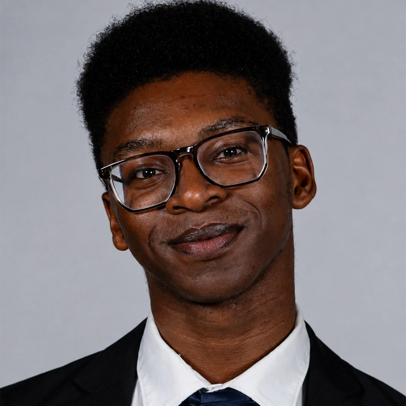 A young Black man with a fade haircut and glasses smiles warmly at the camera. He is wearing a black suit jacket, a crisp white collared shirt, and a dark necktie against a plain grey background.