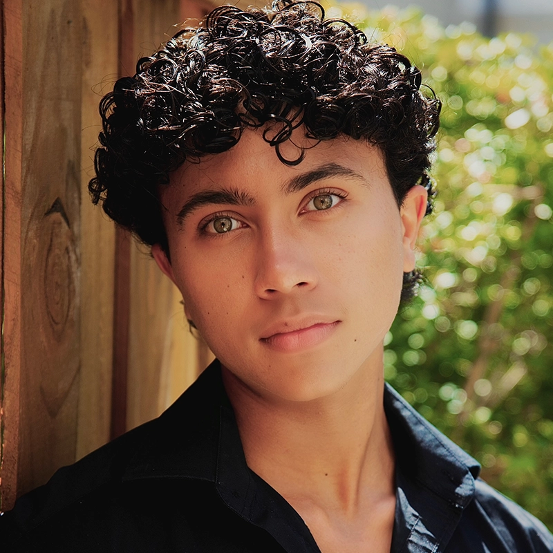 Ryan Bagnolati-Camargo, a young person with curly dark hair and light-colored eyes, looks directly at the camera. They wear a black collared shirt and are positioned outdoors against a blurred background of a wooden fence and green foliage.