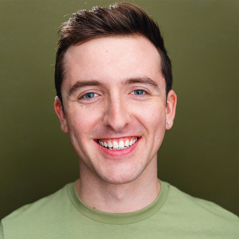 A headshot of a smiling young man with short brown hair, light skin, and blue eyes. He is wearing a light green crew-neck t-shirt against a solid olive-green background.