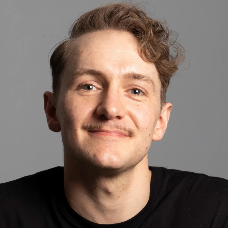 A headshot of a person with wavy light brown hair and a thin mustache, smiling gently at the camera against a plain grey background.